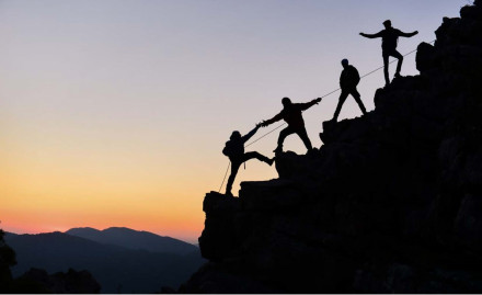 Four people transcending a steep mountain ridge helping each other